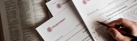 Hand writing on official documents with a pencil, next to an open book with text.