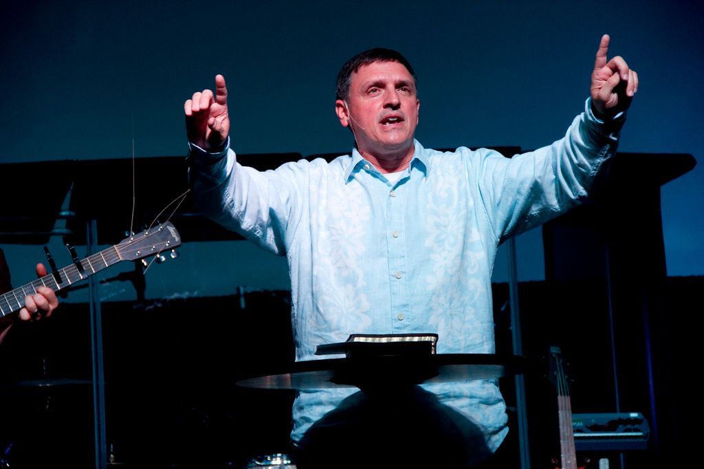 A man is standing at a podium with his arms outstretched and a guitar in the background.
