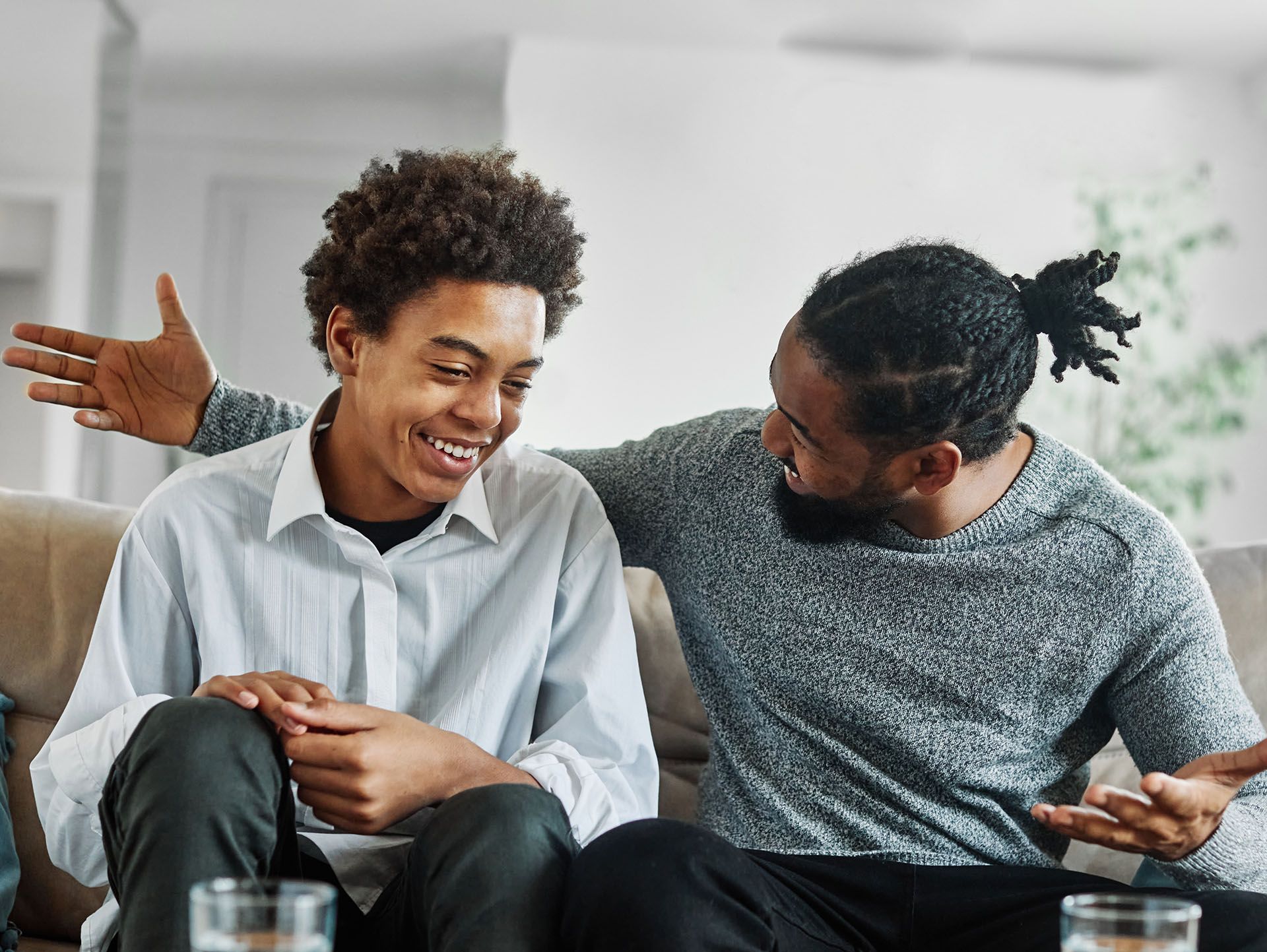 Father with arm around son on a couch, smiling and talking. Indoor setting.