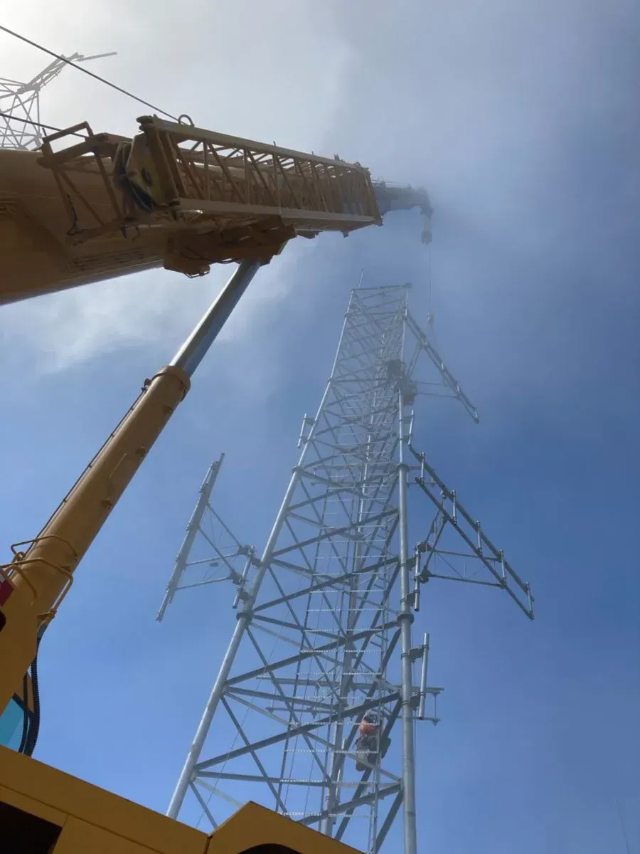 A crane assembling a tall, metal power line tower against a bright blue sky.