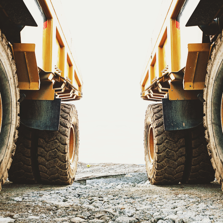 Two yellow dump trucks, side by side, back tires in focus on dirt and rock ground.