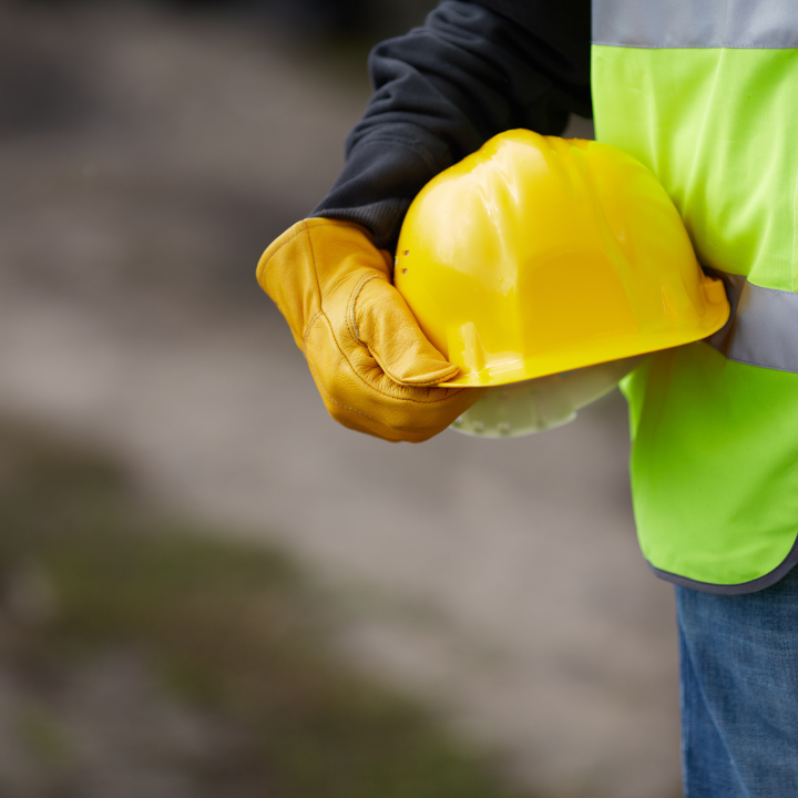 Worker in safety vest holds yellow hard hat, right hand in yellow glove.