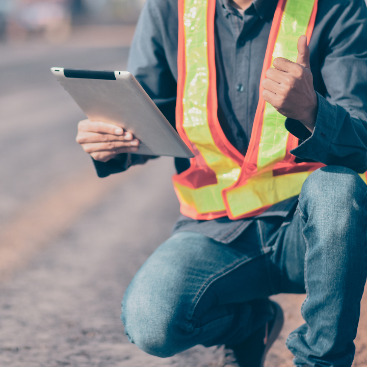 Person kneeling on pavement, holding tablet, wearing safety vest, giving thumbs up.