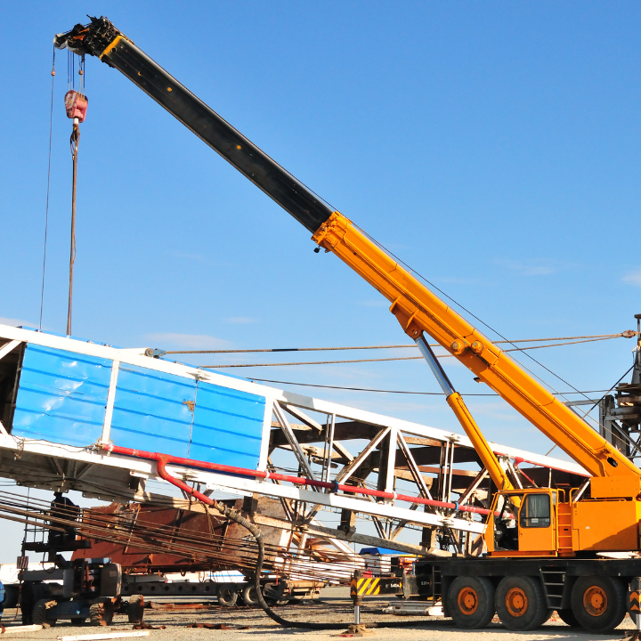 Yellow crane lifting a large, blue-topped structure at a construction site against a blue sky.