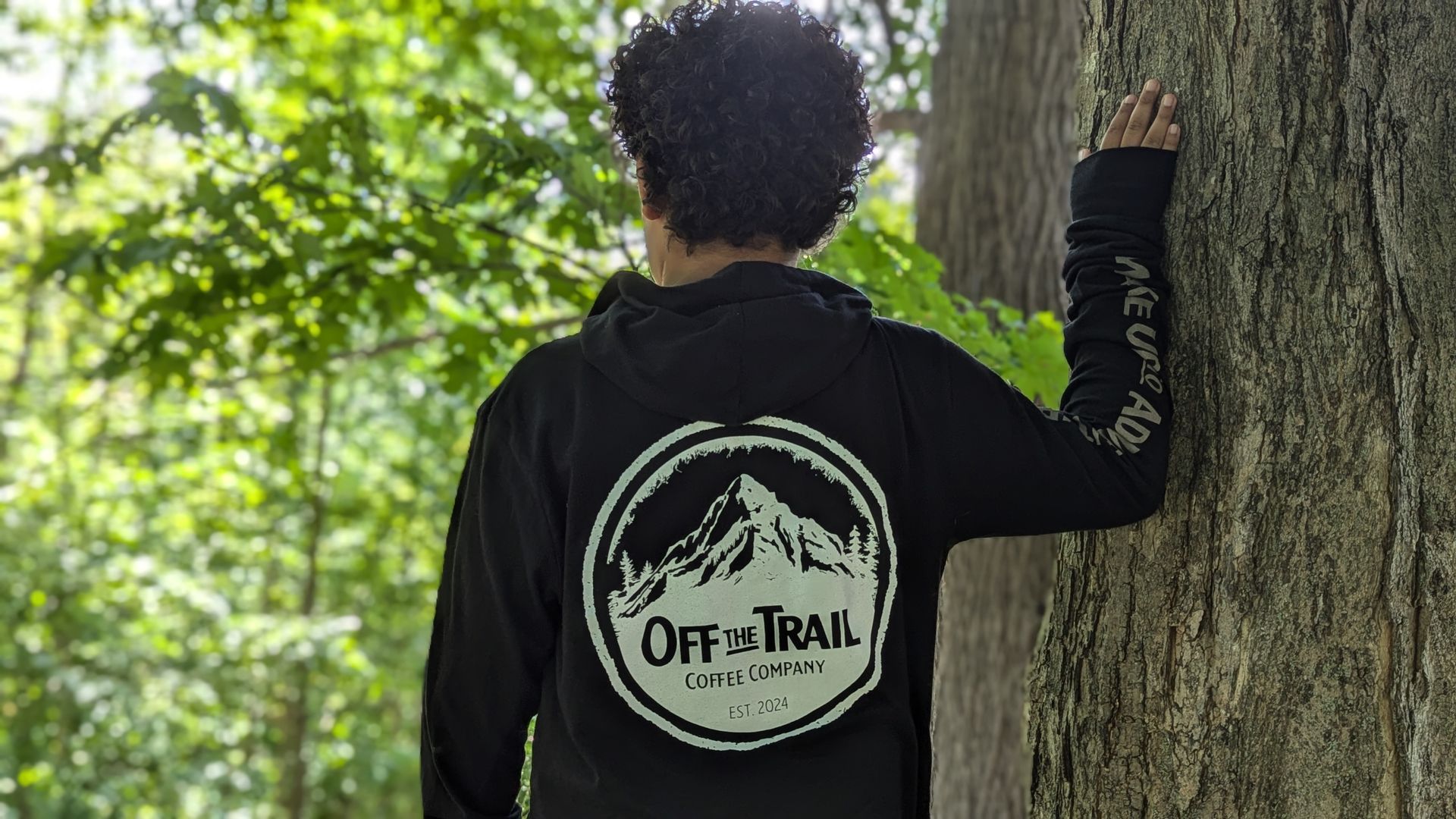 A person wearing a black hoodie is standing next to a tree.