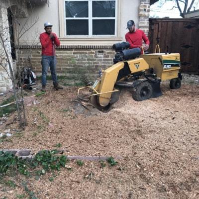Two men are working on a stump grinder in front of a house.