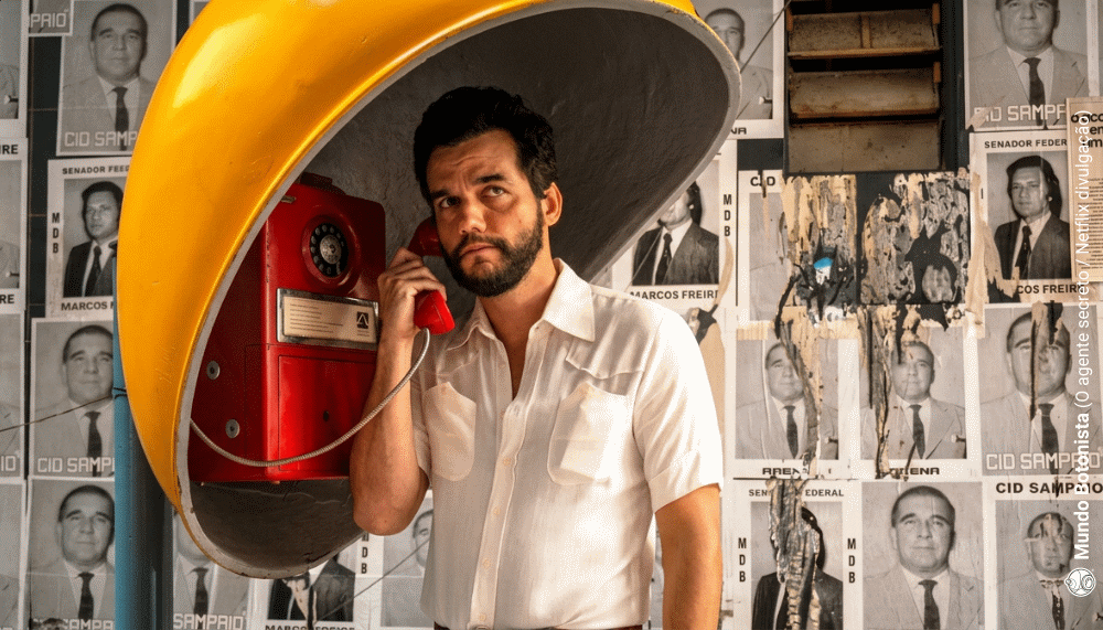 Um homem barbudo, vestindo uma camisa branca, fala ao telefone público amarelo em frente a uma parede coberta de retratos.