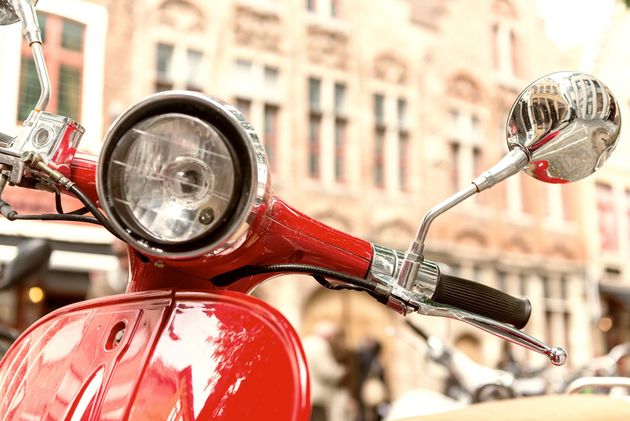 Primo piano di uno scooter vintage rosso con specchietti cromati, parcheggiato in una strada cittadina con edifici sfocati sullo sfondo.