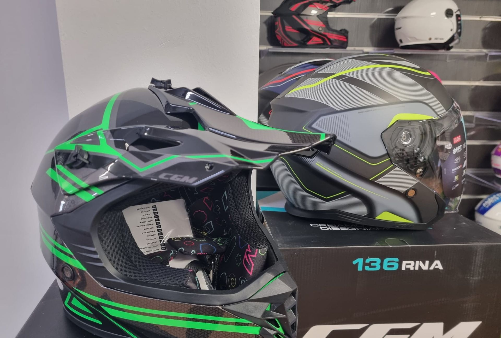 Un casco da motocross nero e verde accanto a un casco grigio su uno scaffale, con un casco bianco sullo sfondo.