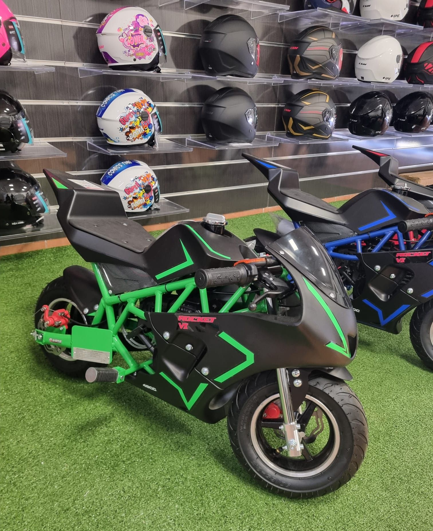 Due piccole motociclette sportive, verde e nera nella parte anteriore, esposte in uno showroom insieme a caschi sugli scaffali.