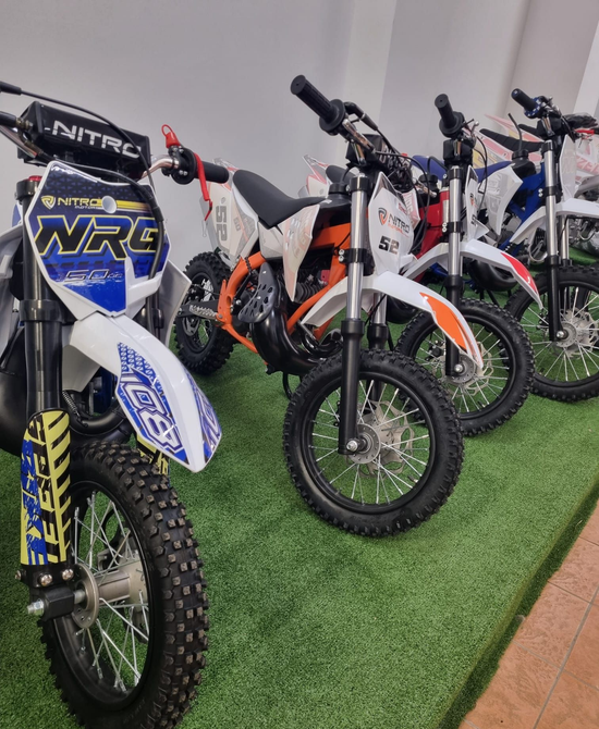 Una fila di moto da cross su un campo in erba sintetica al coperto, tra cui una moto anteriore blu e bianca e moto arancioni in stile KTM.