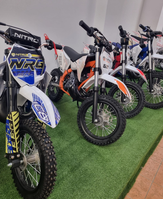 Una fila di moto da cross al coperto su un prato artificiale, con disegni blu, arancioni e bianchi ben visibili.