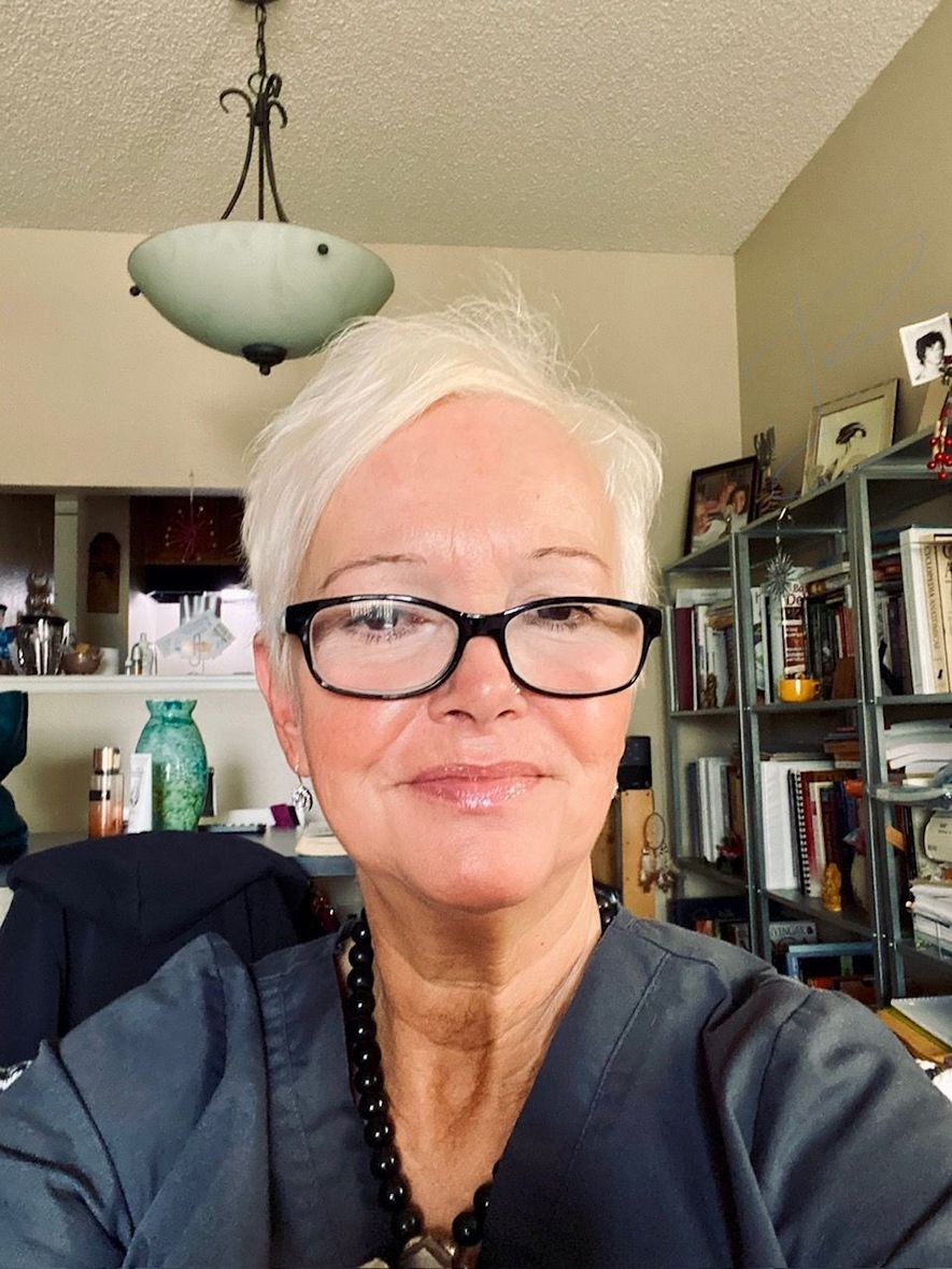 Woman with short white hair and glasses smiles; indoors with desk and shelves visible.