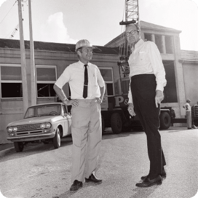 Two men in work attire stand near a truck and building, one with a hard hat, discussing construction.