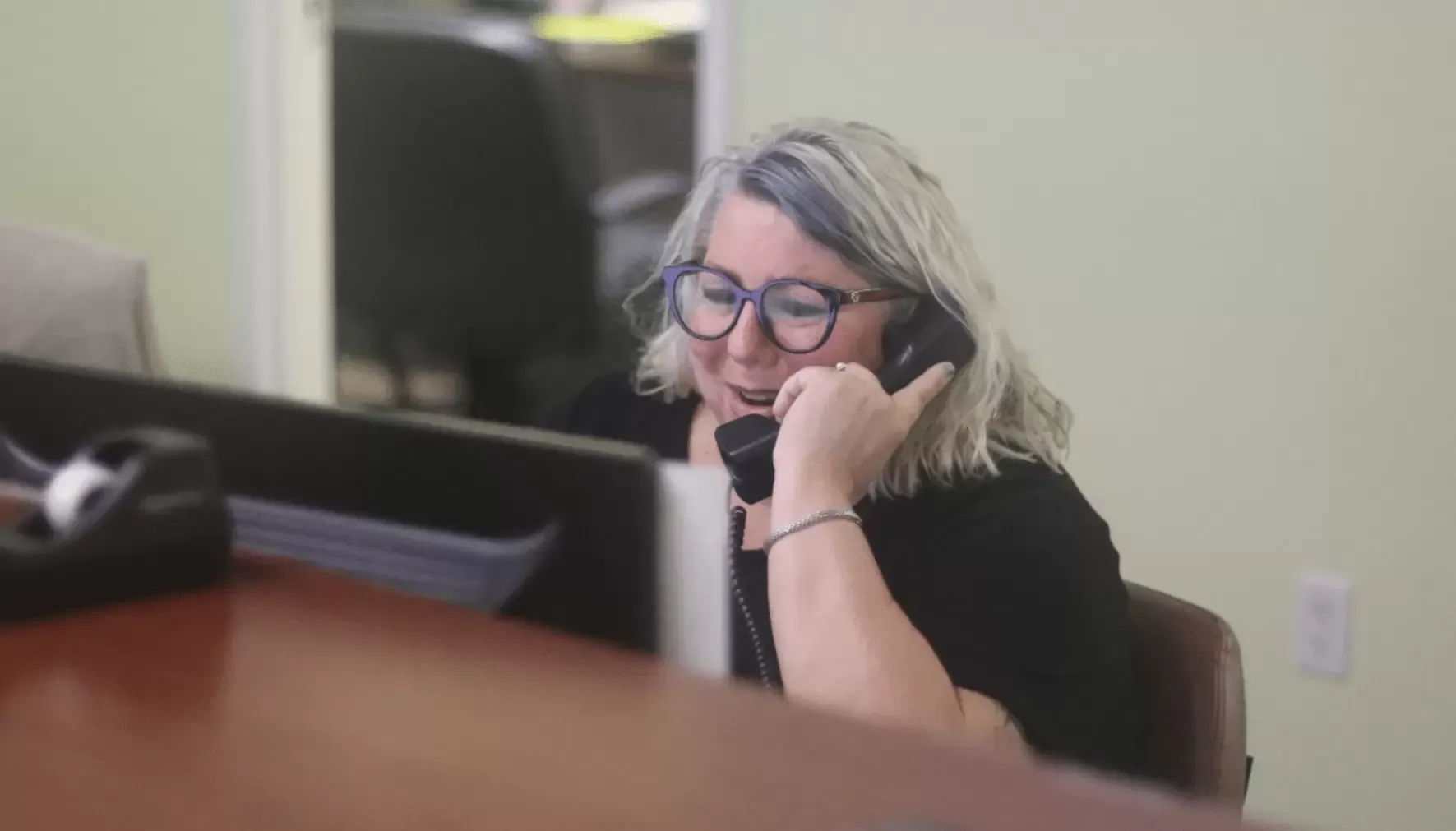 Woman with glasses smiling while on the phone at a desk.