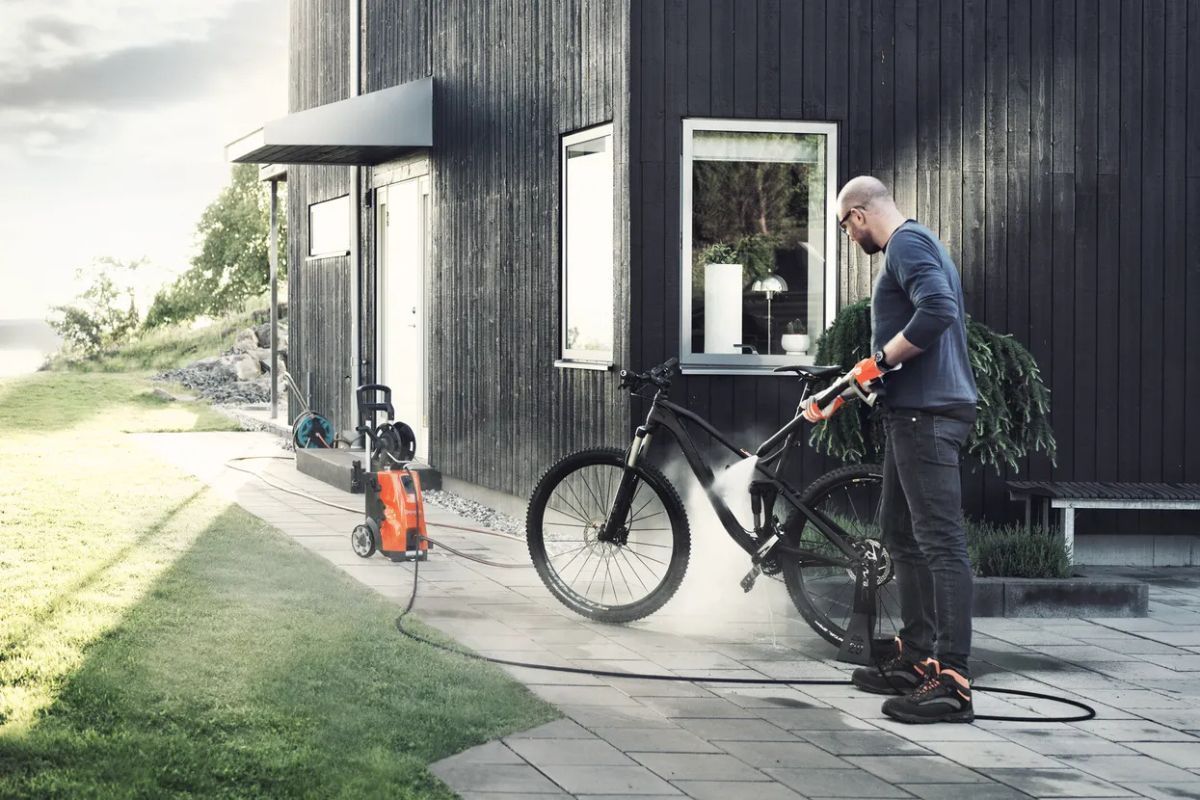 Man washing a black mountain bike with a pressure washer on a patio next to a dark house.