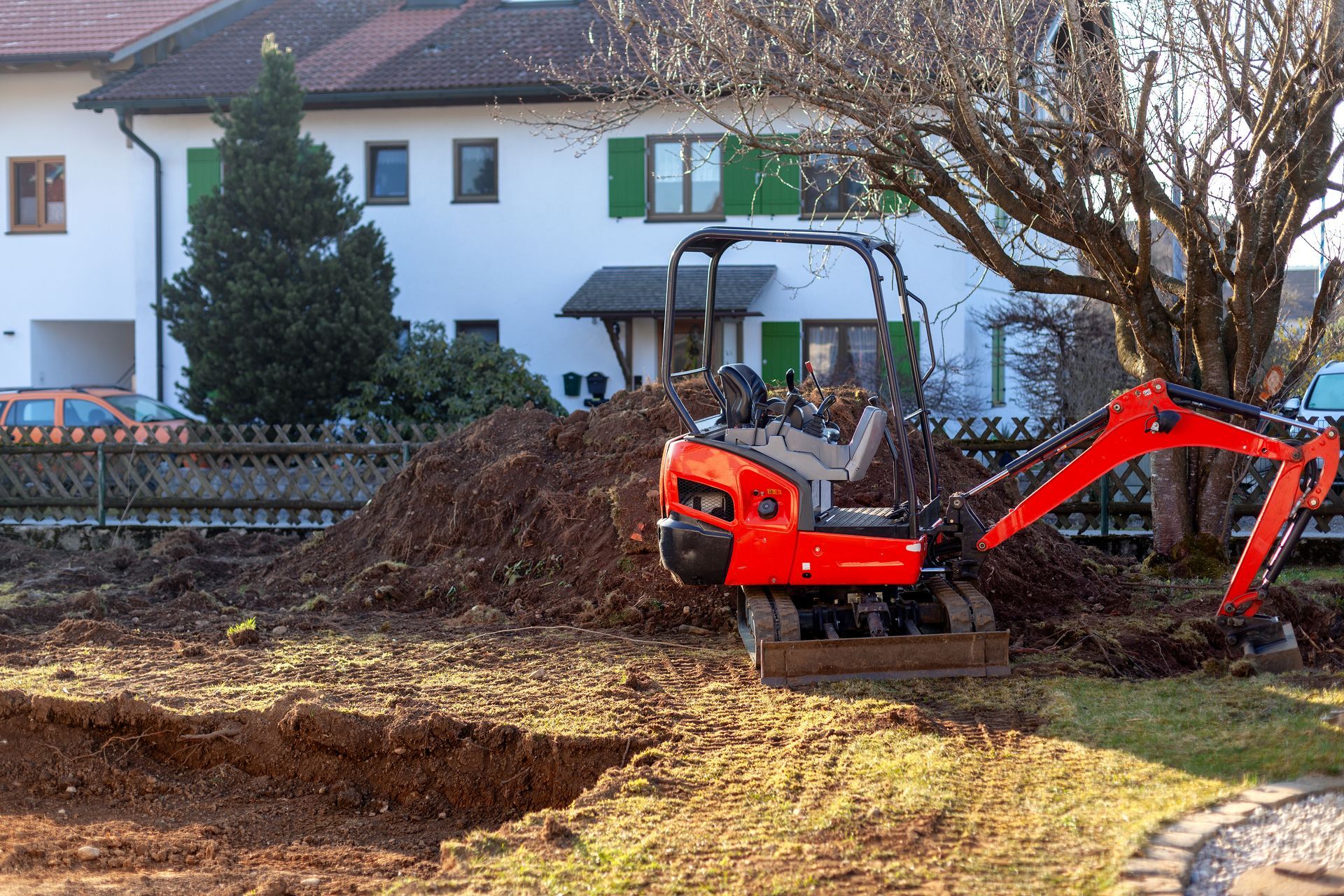 Small red excavator digging in a yard in front of a white house with green shutters.