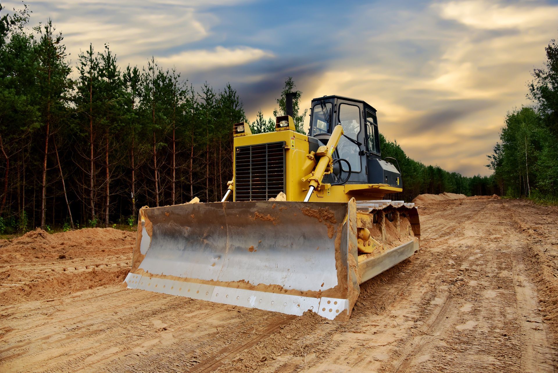 Yellow bulldozer clearing a dirt road in a forest, under a cloudy sky.