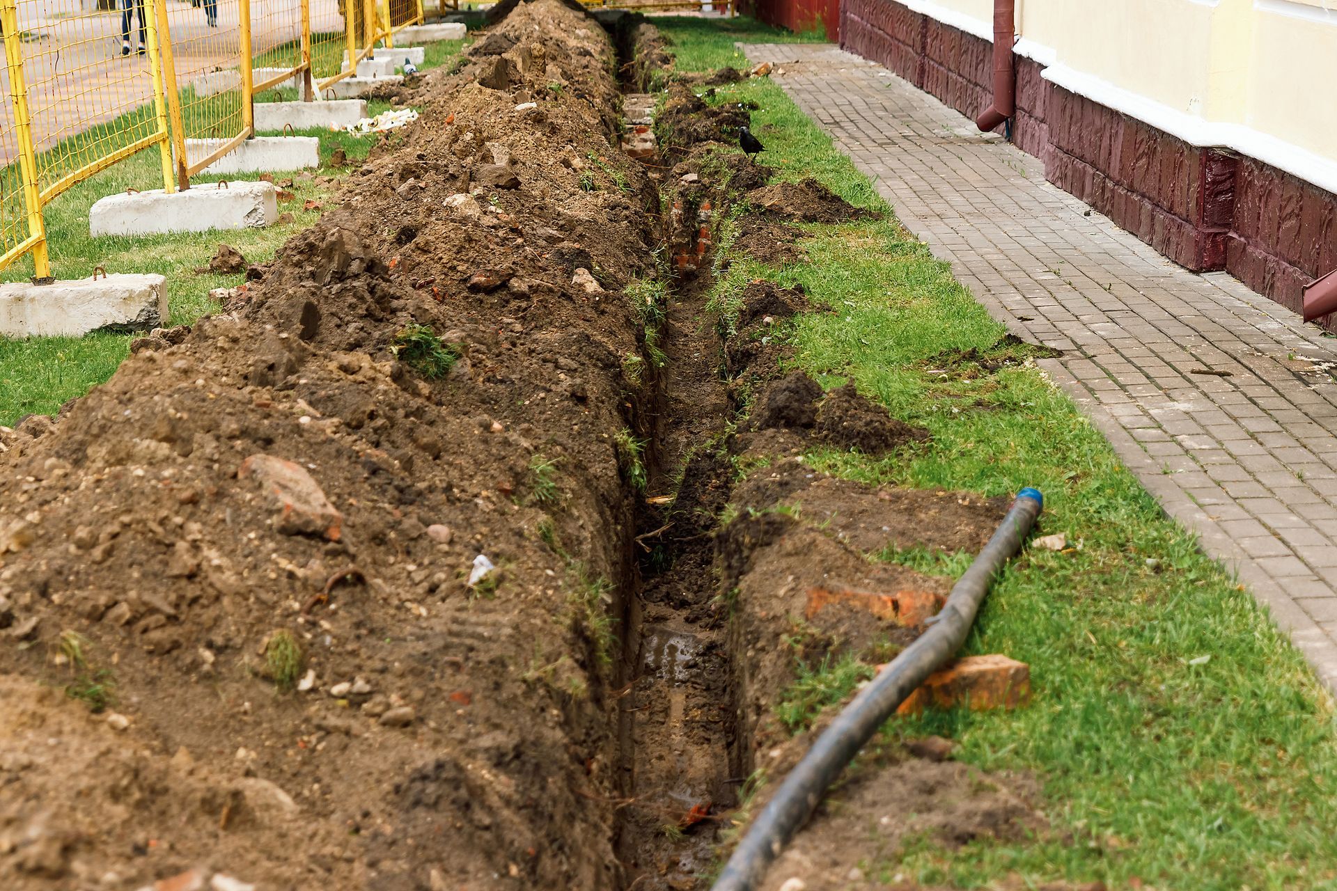 Trench dug in the ground next to a building and sidewalk, containing a black pipe.