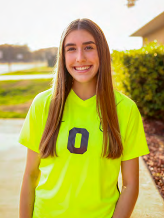 A young woman wearing a neon yellow soccer jersey is standing on a sidewalk.