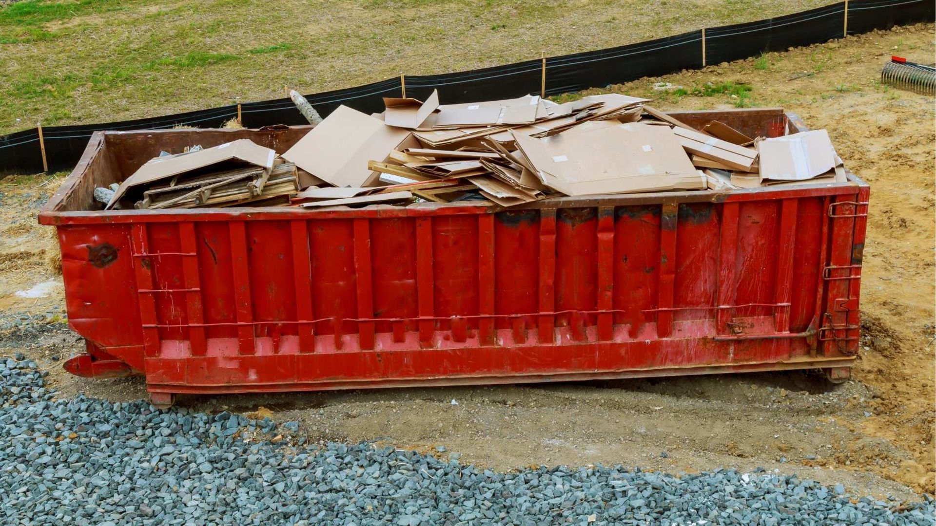 Red dumpster filled with cardboard debris on a gravel surface.