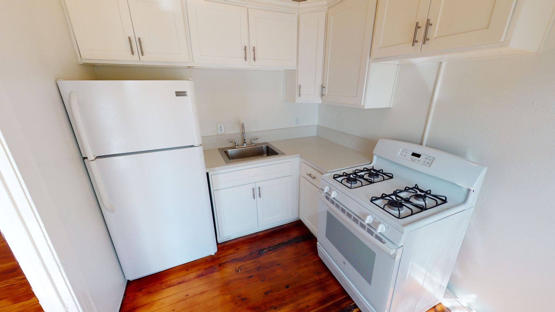 Small, all-white kitchen with a fridge, sink, cabinets, and a gas stove on wooden flooring.