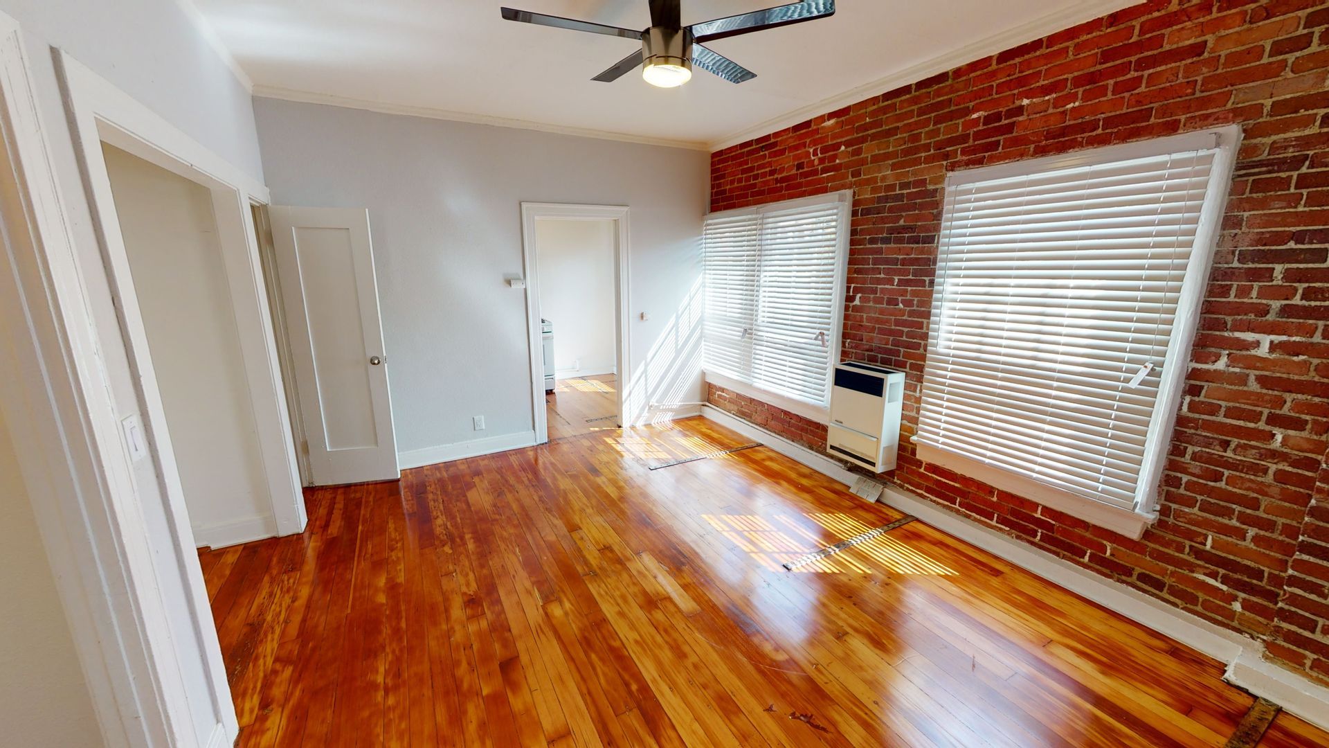 Empty room with wood floors, brick wall, and blinds. Sunlight streams through.