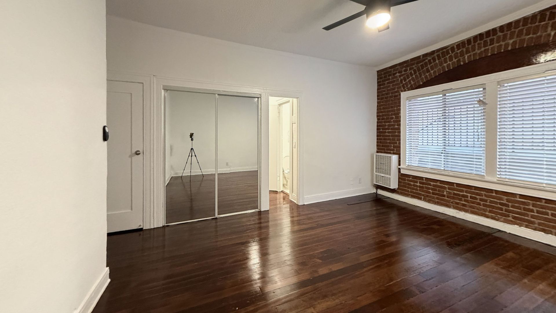 Empty room with wooden floors, brick wall, window, and a door to another room.