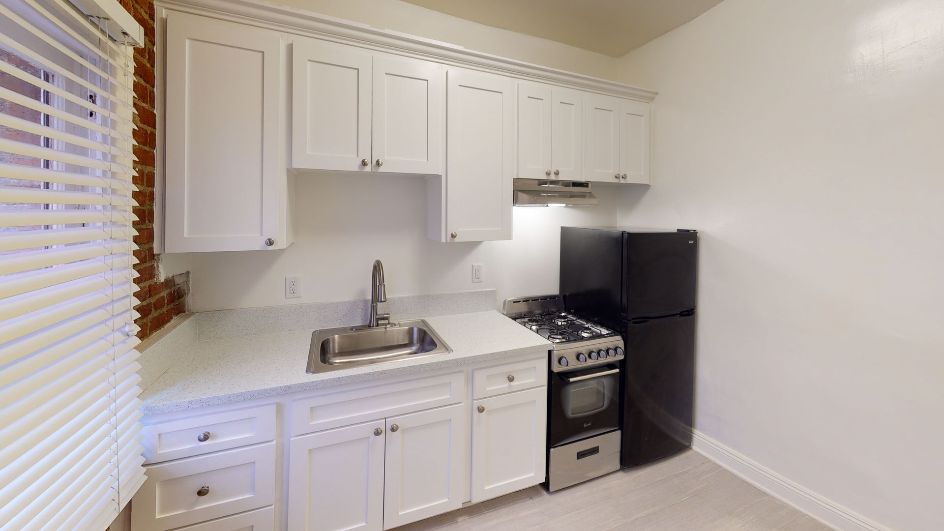Small white kitchen with cabinets, sink, stove, and black refrigerator. Window with blinds on the left.