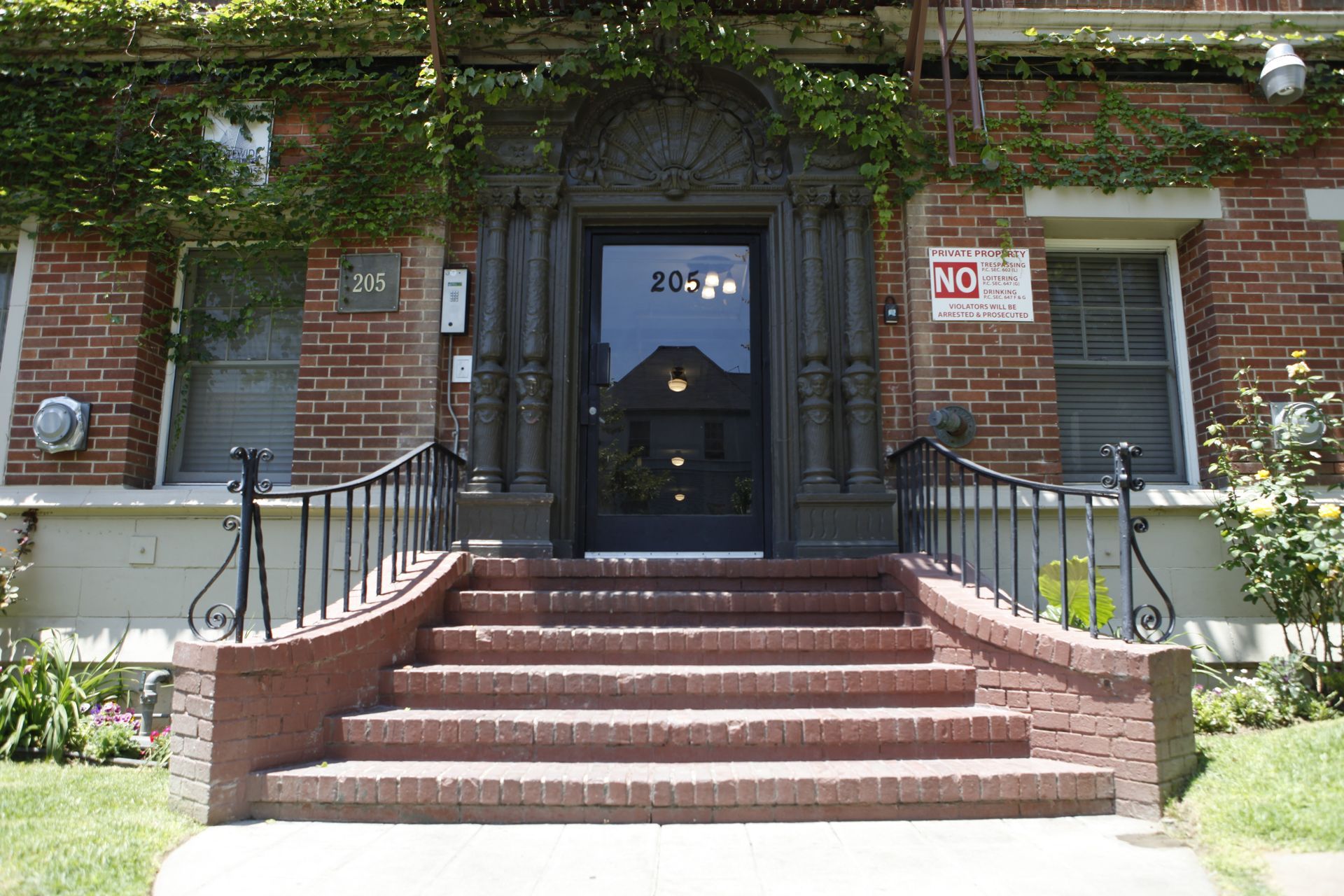 Brick building entrance with stairs, black door, and ornate archway covered in vines.