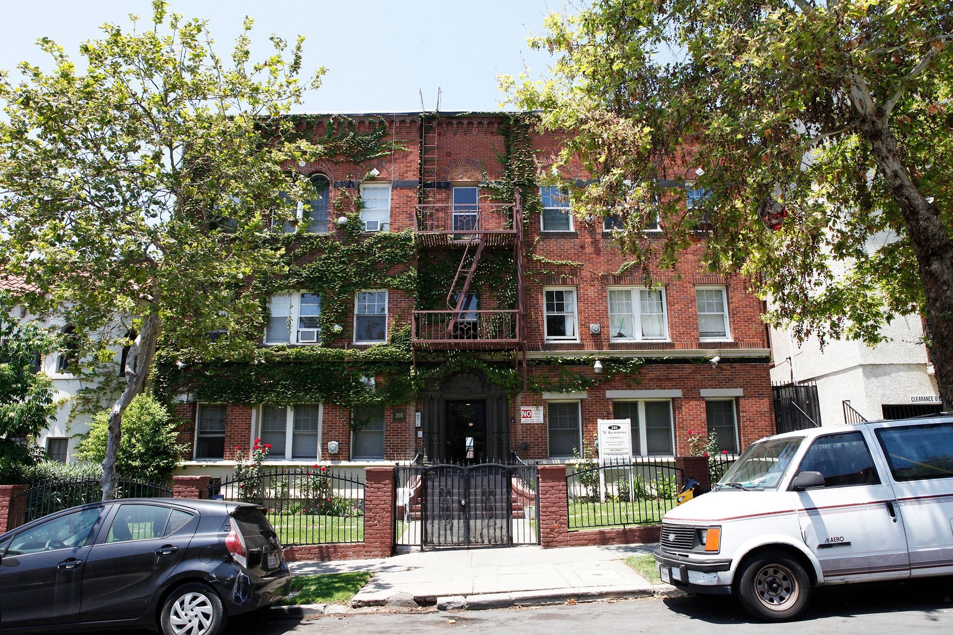 Brick apartment building with green vines, wrought iron gate, and parked vehicles.