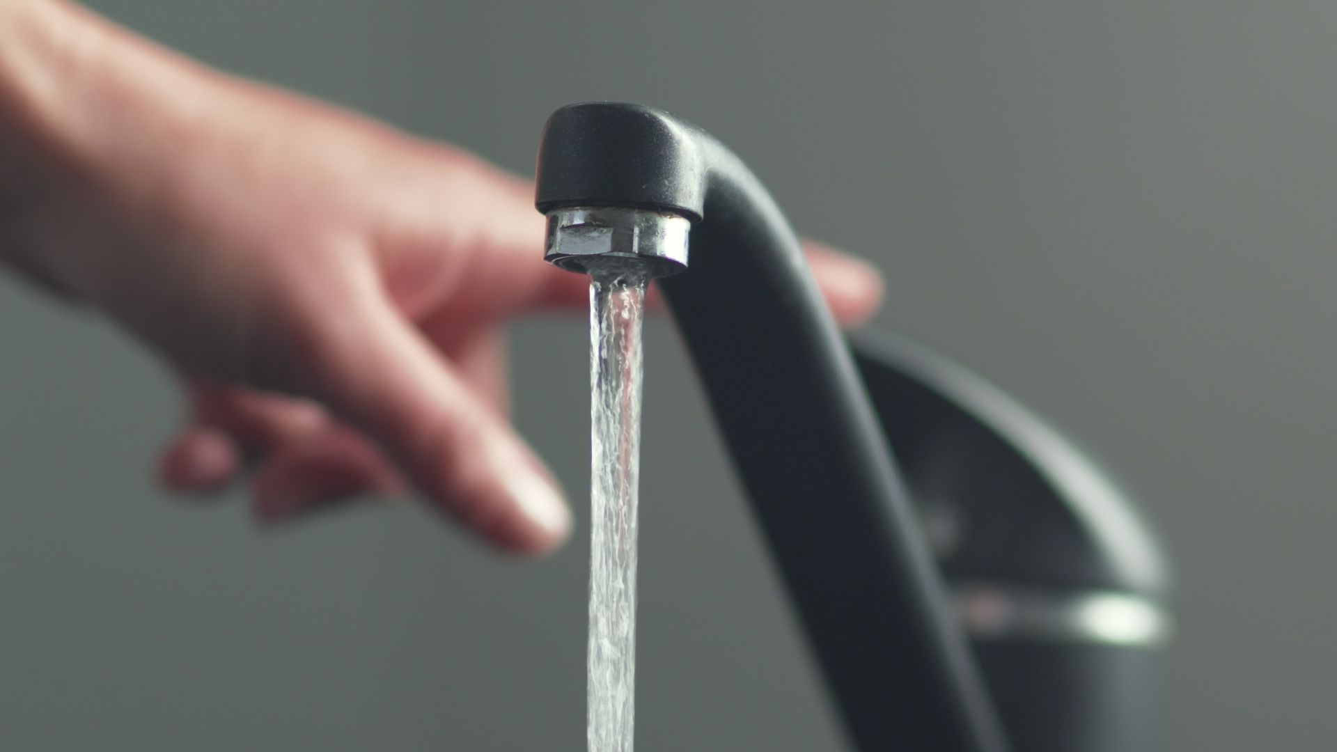 Close-up of water running from a black faucet with a hand reaching toward it.