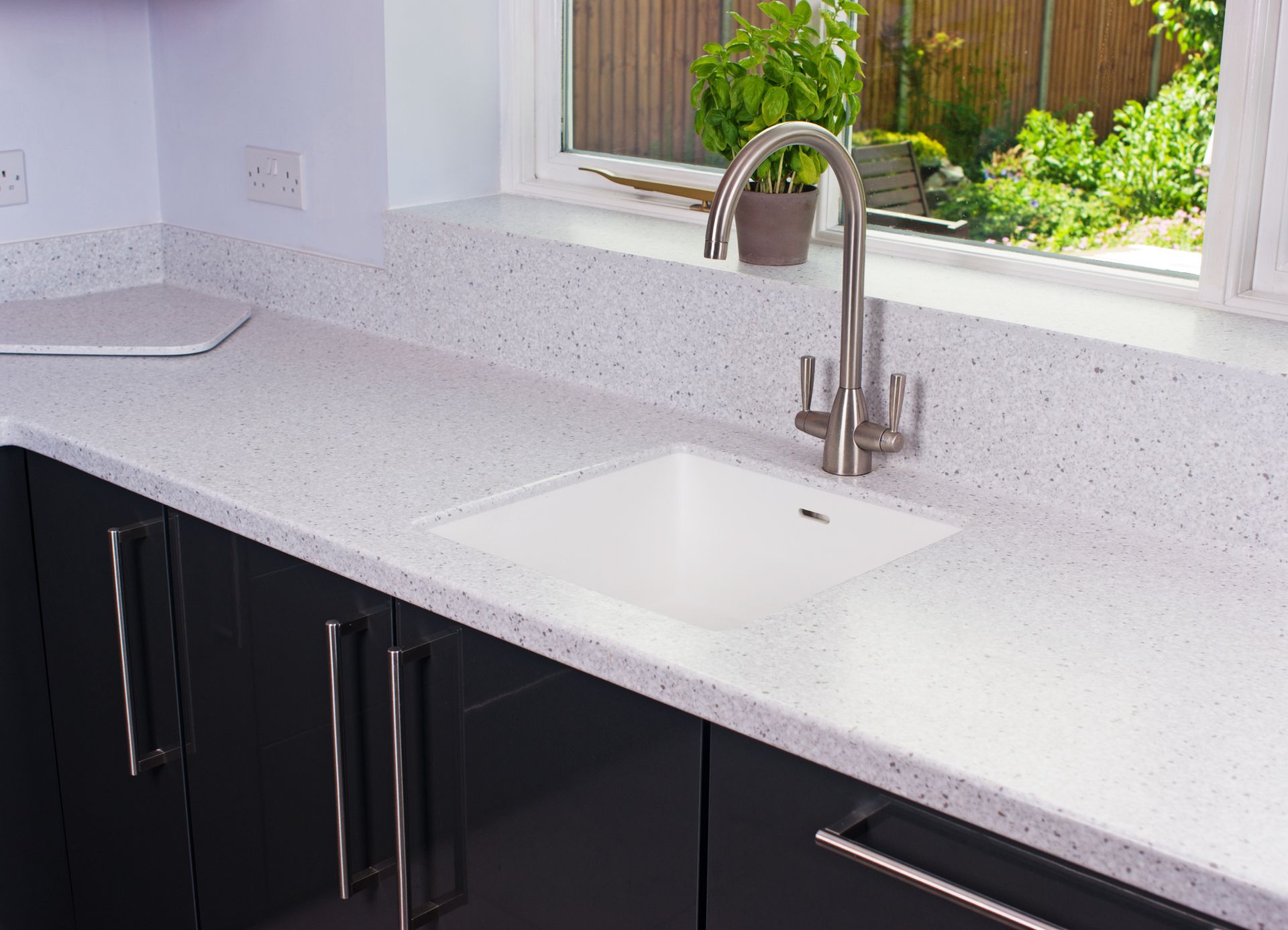 Kitchen with a white countertop, a silver sink and black cupboards. A window opens to a yard