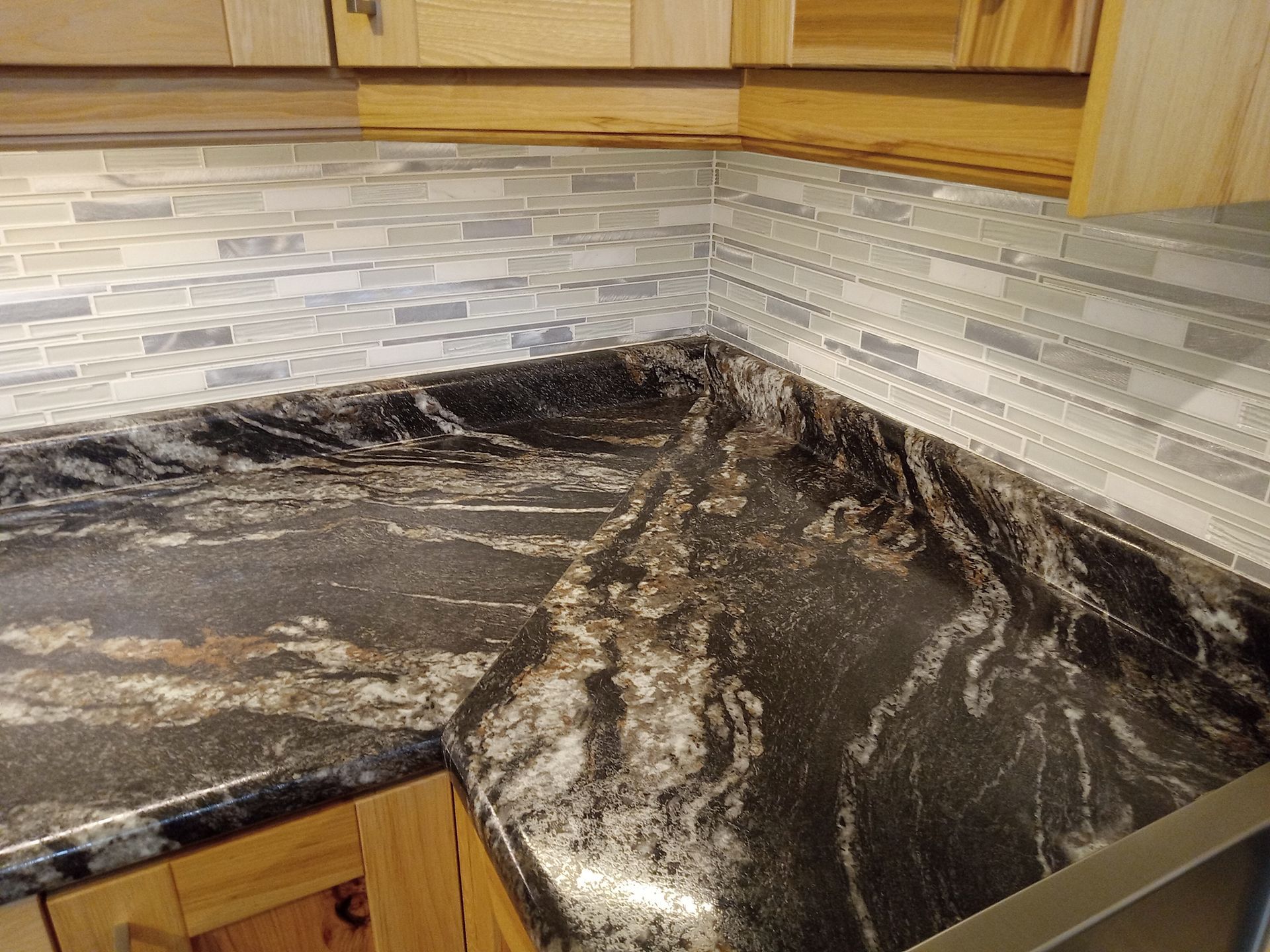 A kitchen corner with a dark granite countertop, light wood cabinets, and a gray tile backsplash