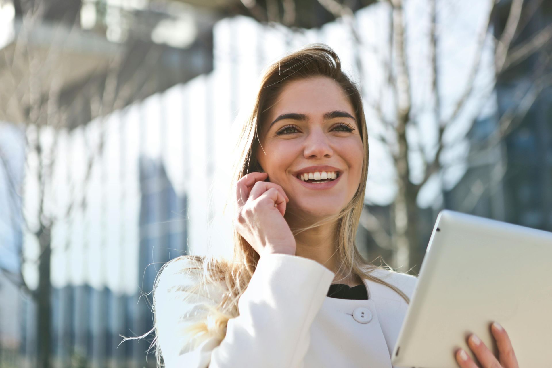 Woman in a white jacket smiling, holding tablet, talking on the phone outside a building.