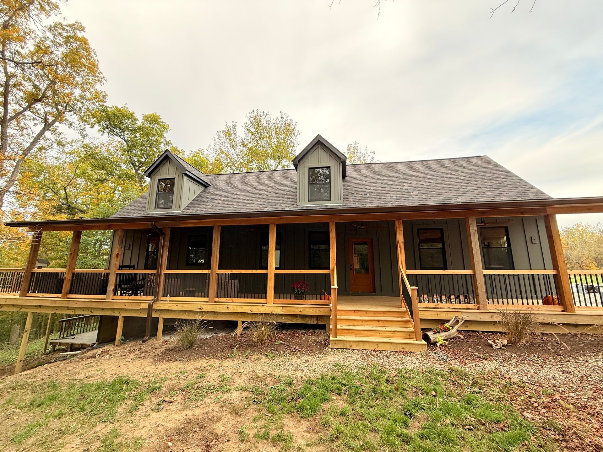 Cabin with wraparound porch and two dormers; gray siding, brown trim, and steps leading to a wooden door.