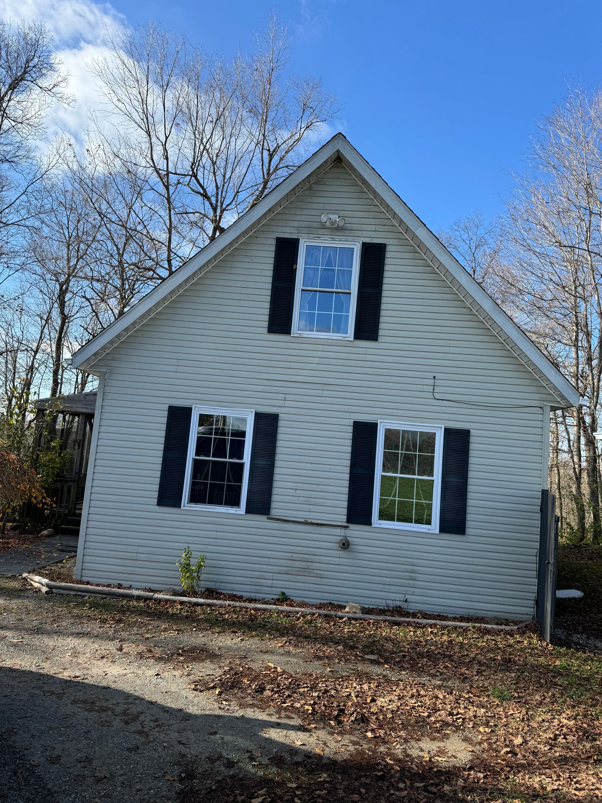 Beige house with black shutters, two windows on each level, surrounded by trees under a blue sky.