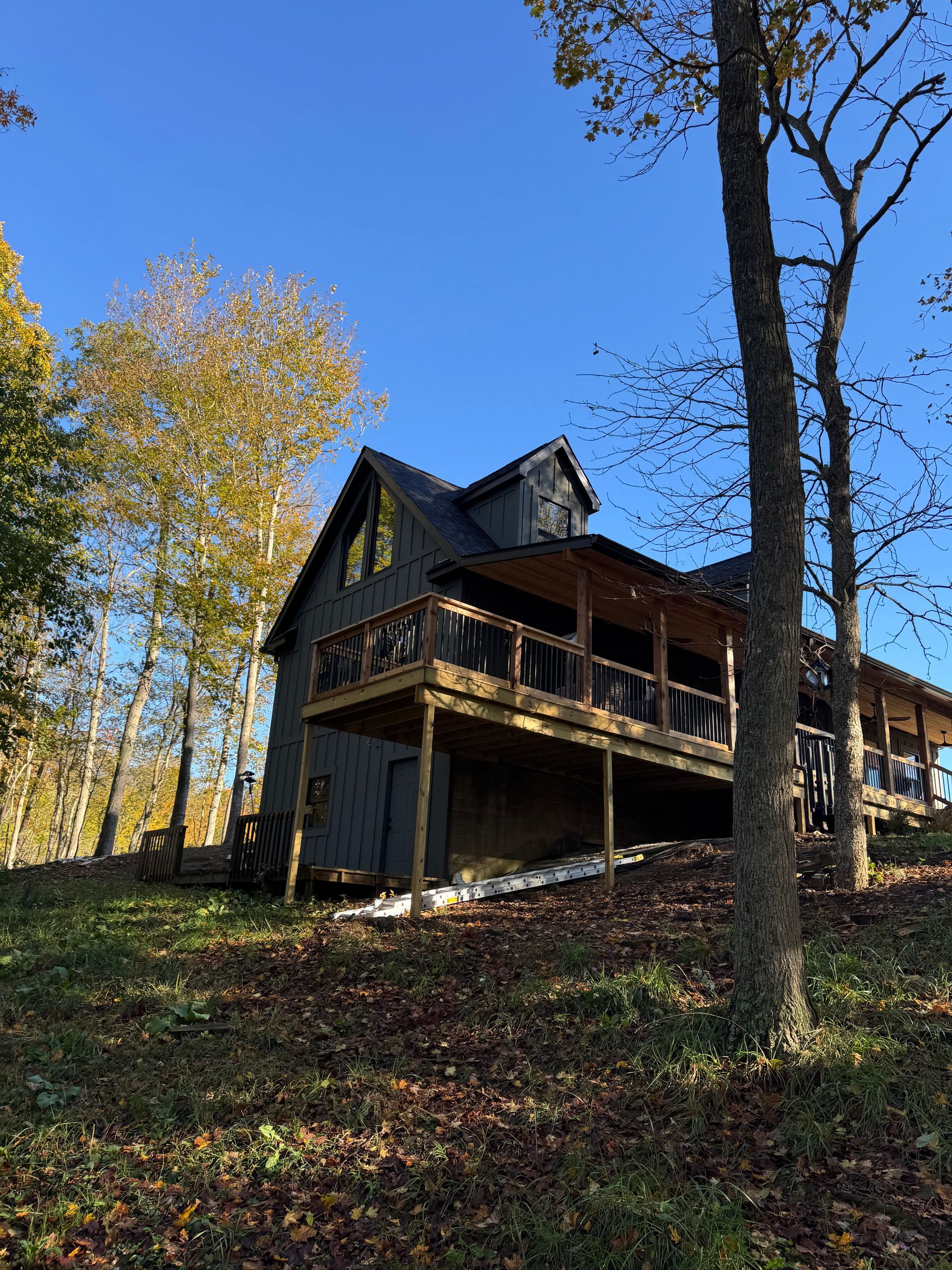 Wooden cabin with a dark gray exterior and a wraparound deck on a hillside under a blue sky.