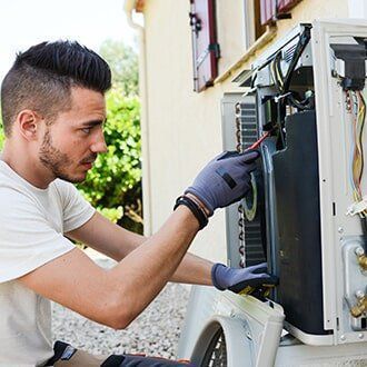 A technician in grey gloves repairs an air conditioning unit outside a building. He is focused and using a tool.