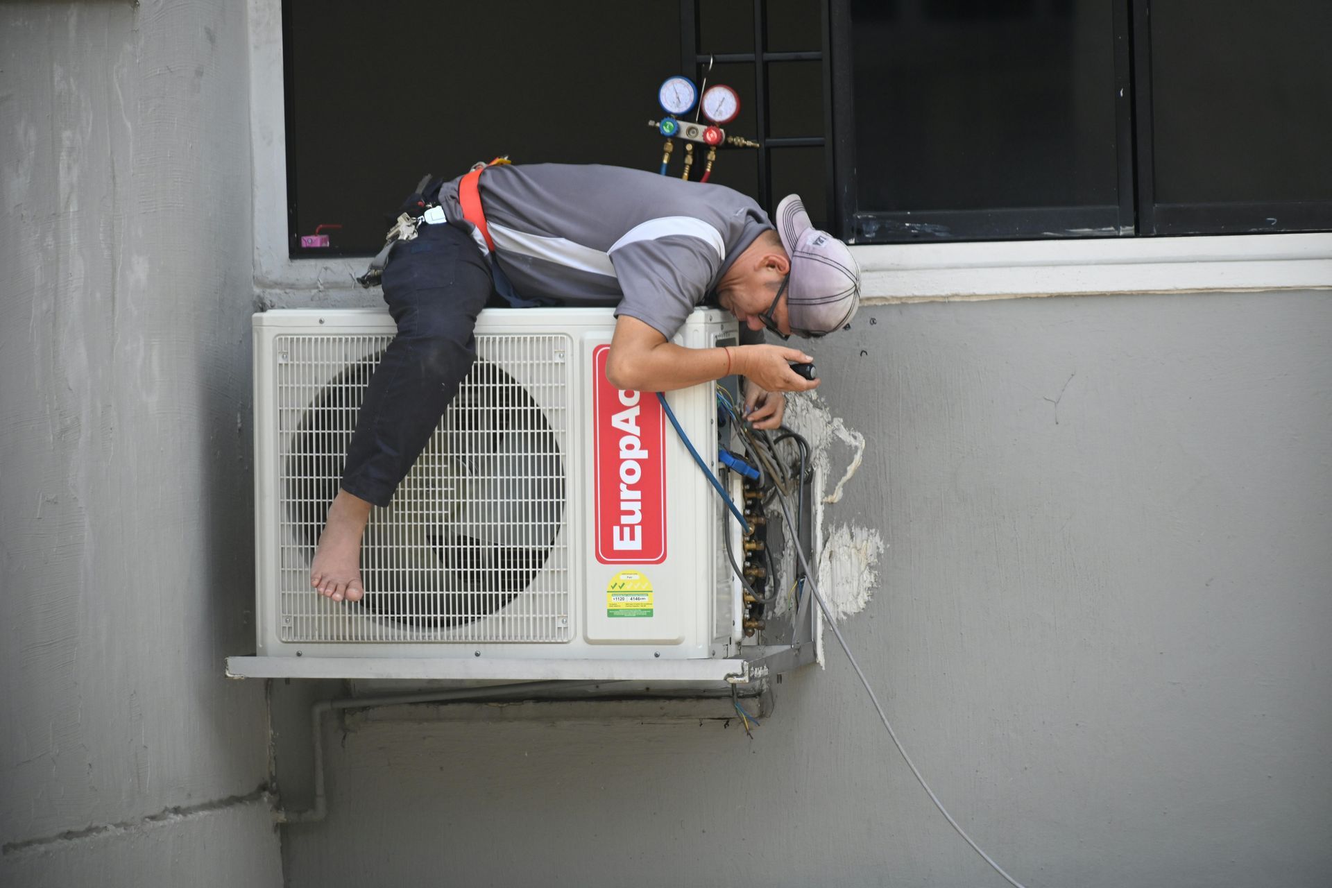 A man is working on an outdoor air conditioning unit, perched precariously with his legs dangling. He is wearing a grey shirt and a cap.