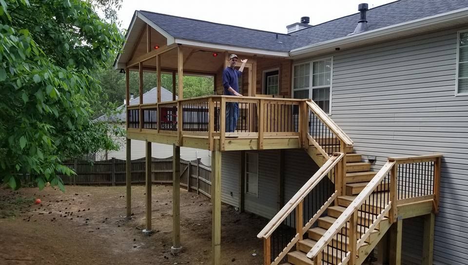 A person stands on a wooden deck with a covered porch area attached to the back of a grey-sided house with outdoor stairs.