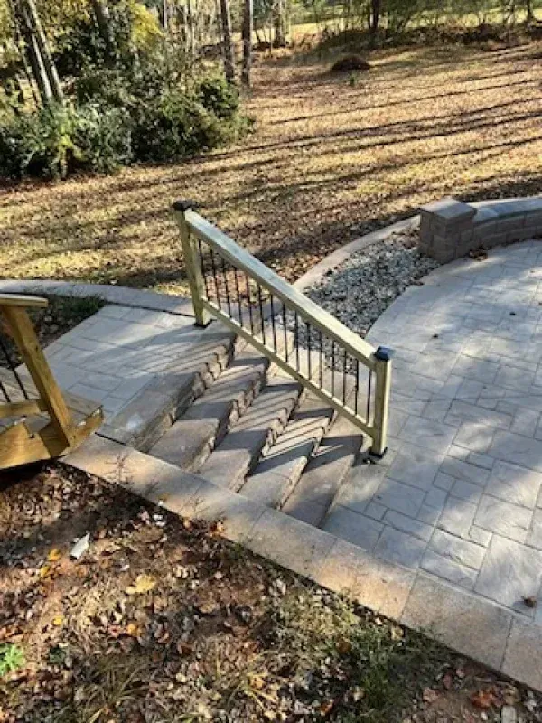 Outdoor concrete stairs with a tan handrail and black balusters descending from a stone patio to a grassy lawn area.