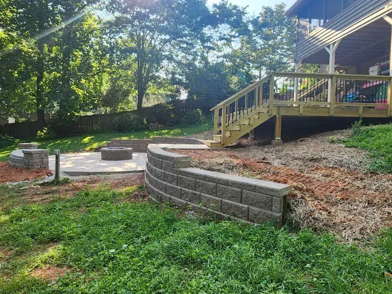 A backyard features a stone fire pit area, a curved retaining wall, and a wooden deck with stairs leading to a house.