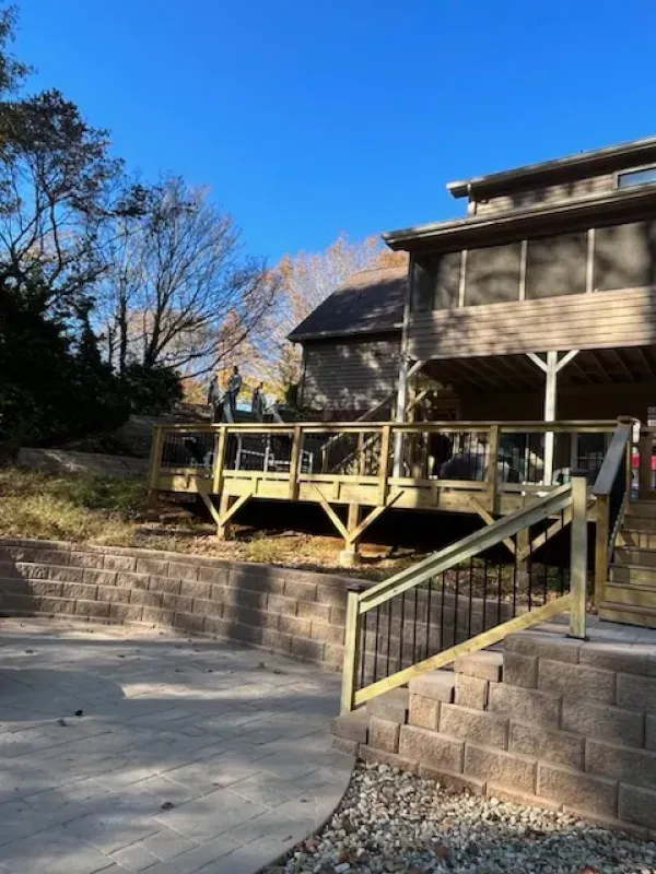 A wooden deck with a staircase overlooks a multi-tiered stone patio and retaining wall under a clear blue sky.