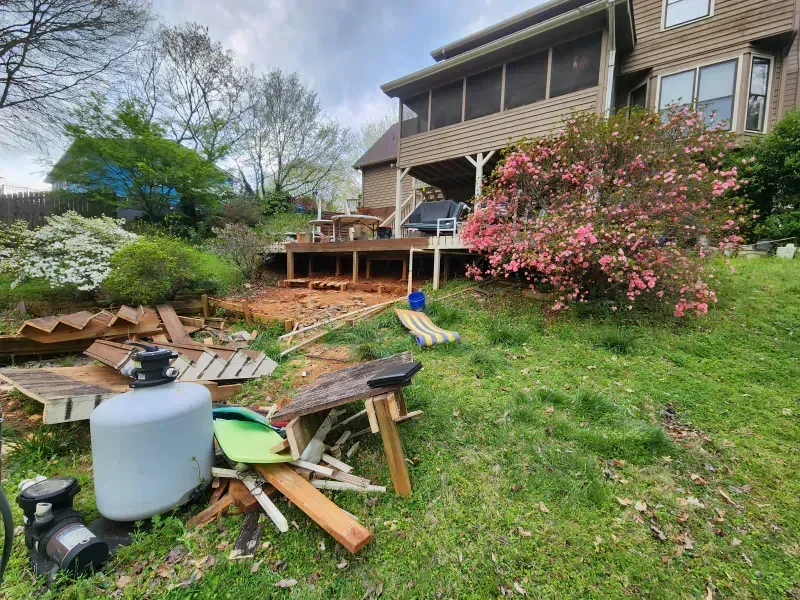 A grassy backyard showing a house with a wooden deck under construction, surrounded by piles of discarded lumber and debris.