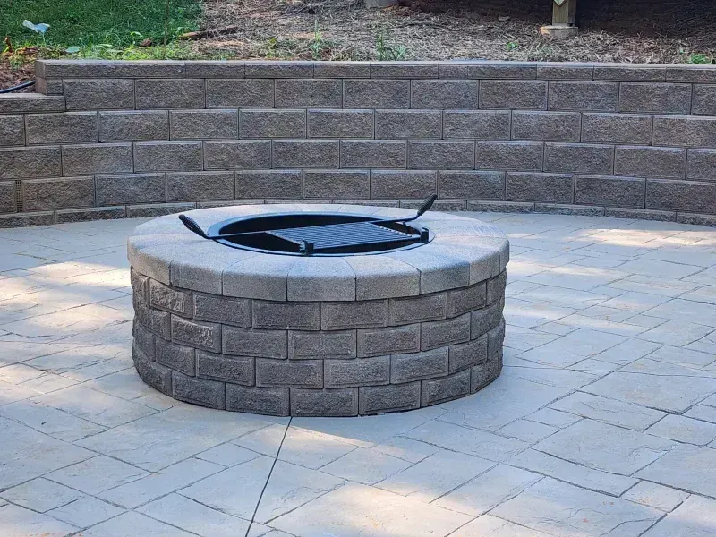 A circular stone fire pit sits on a light-colored paver patio in front of a curved stone retaining wall.