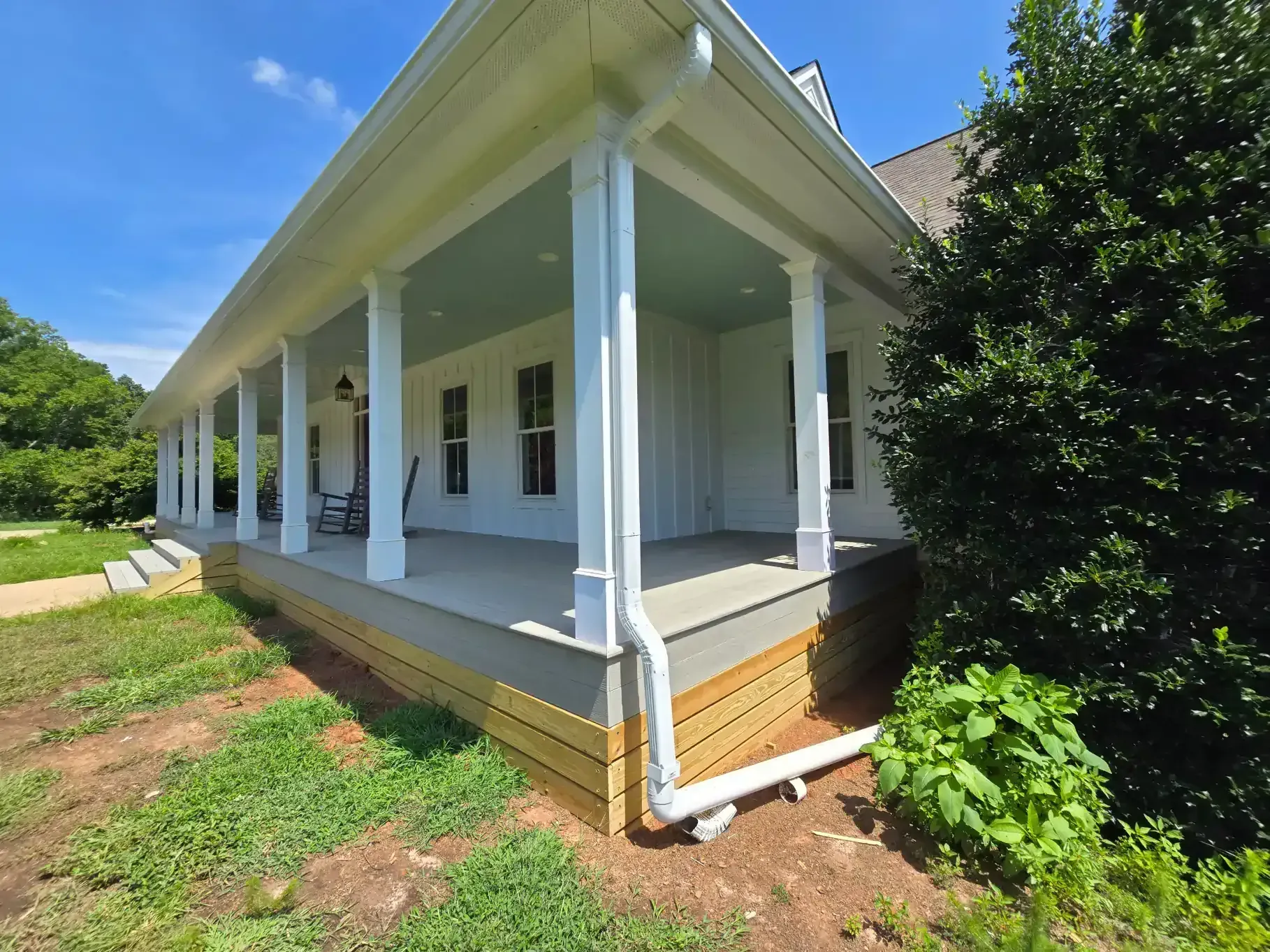 White farmhouse porch with wooden skirting, white columns, and a light blue ceiling under a clear blue sky.
