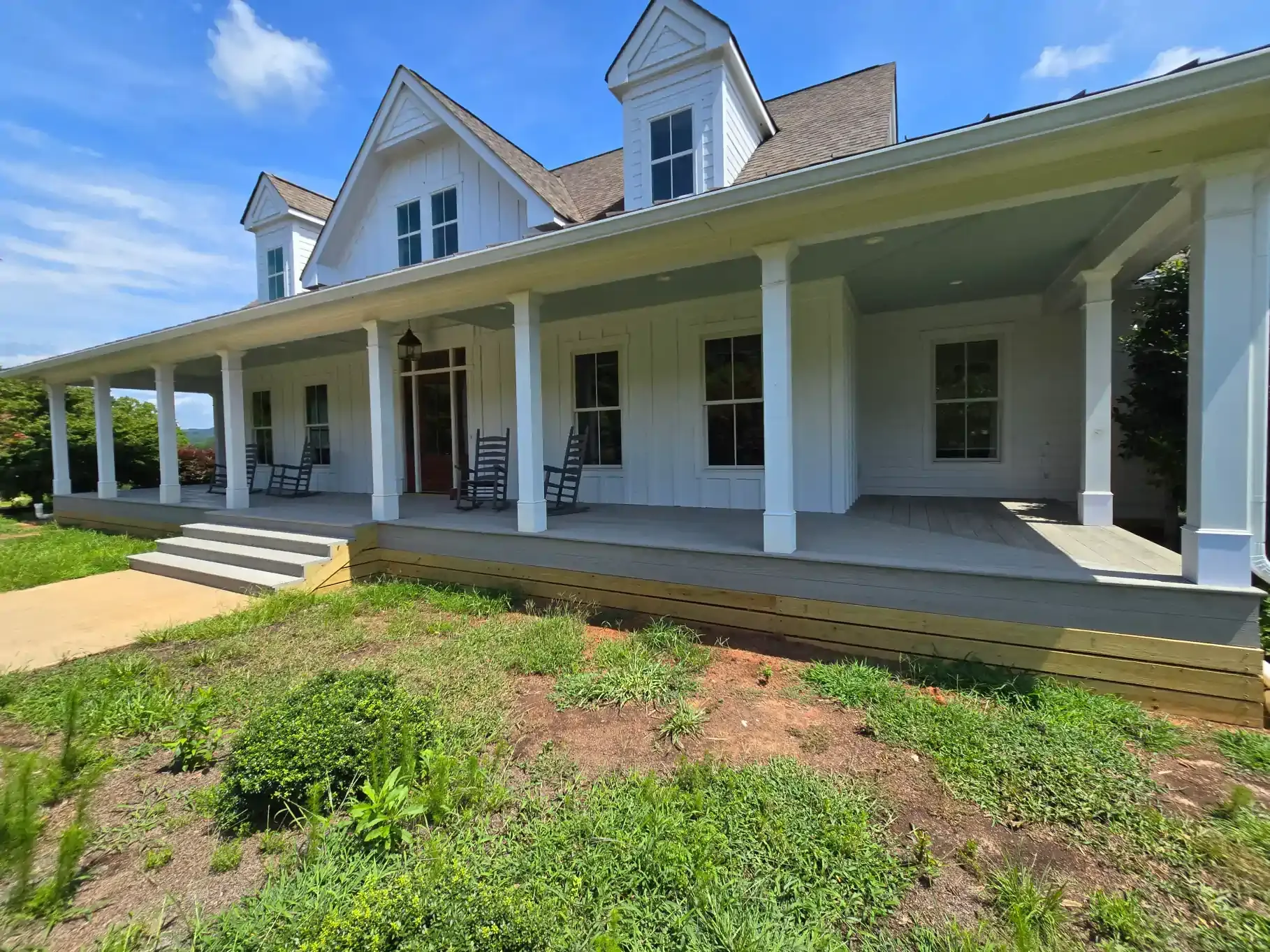 A bright white house with a wide, covered front porch, columns, and a grassy front yard under a clear blue sky.