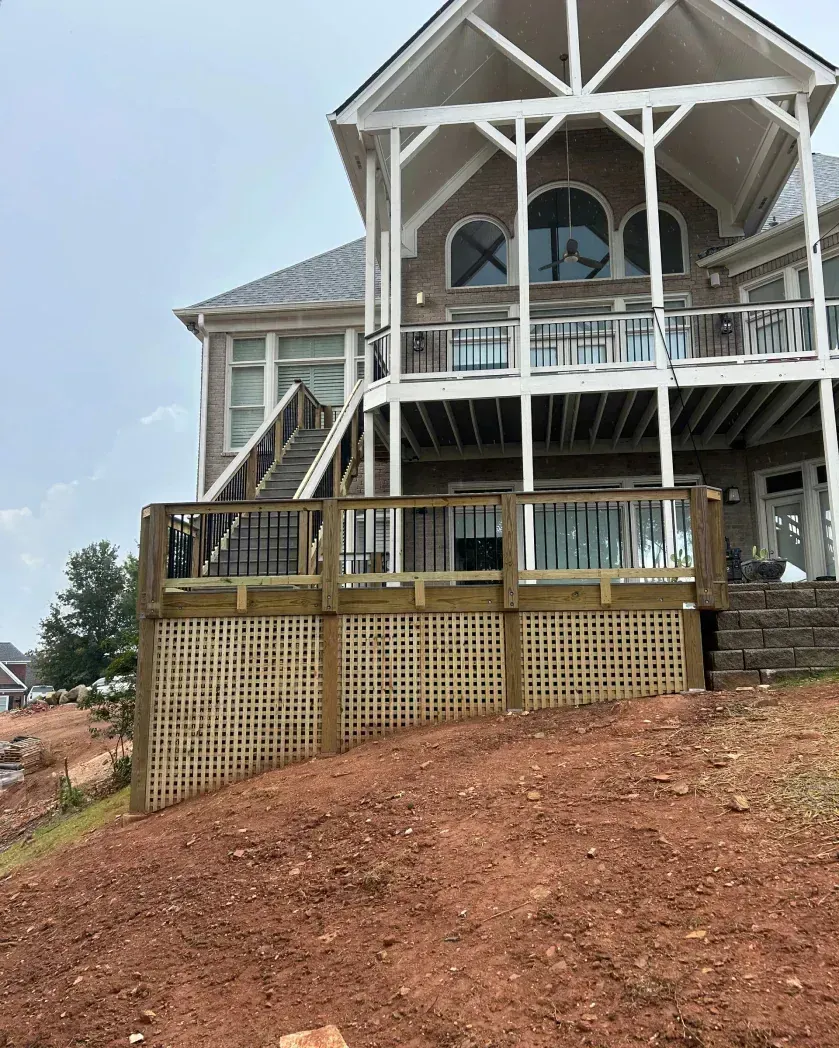 A large house with a multi-level deck and lattice skirting, overlooking a sloped, red-dirt yard.
