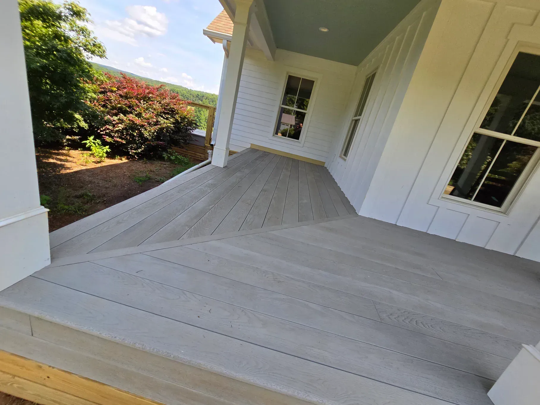 A wide, gray-planked porch wraps around the corner of a white house with a blue-painted ceiling under the eaves.